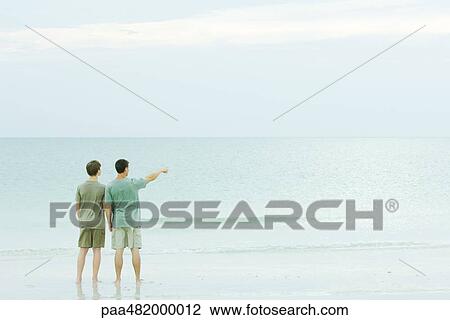 Stock Image - Father and son standing together at the beach, looking at view, one pointing. Fotosearch