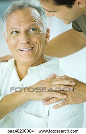 Stock Image - Mature man and adult son smiling at each other, son's hand on father's shoulder, cropped view. Fotosearch