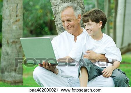 Mature man holding grandson on lap, both looking at laptop computer and smiling View Large Photo Image Stock Photo - Mature man holding grandson on lap, both looking at laptop computer and smiling. Fotosearch