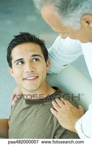Stock Photo - Mature man leaning over, placing hands on son's shoulders, both smiling at each other. Fotosearch