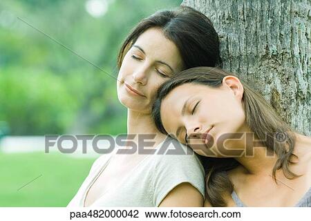 Stock Image - Mother and teenage daughter leaning against tree trunk with eyes closed, girl's head on woman's shoulder. Fotosearch