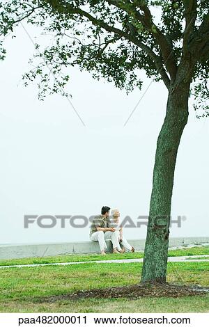 Stock Image - Senior woman and adult son sitting together outdoors, mid-distance, tree in foreground. Fotosearch