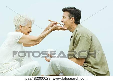 Stock Image - Senior woman feeding adult son takeout food with chopsticks, side view. Fotosearch