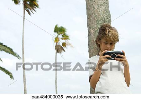 Boy leaning against tree trunk, playing handheld video game View Large Photo Image Stock Photography - Boy leaning against tree trunk, playing handheld video game. Fotosearch
