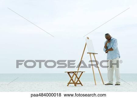 Male painter standing in front of blank canvas at the beach, hand under chin, looking down View Large Photo Image Stock Photo - Male painter standing in front of blank canvas at the beach, hand under chin, looking down. Fotosearch