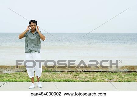 Man standing in front of beach, listening to headphones, hands raised to head View Large Photo Image Stock Image - Man standing in front of beach, listening to headphones, hands raised to head. Fotosearch