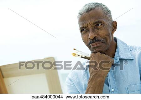 Picture - Senior man holding paint brushes, hand under chin, looking away. Fotosearch