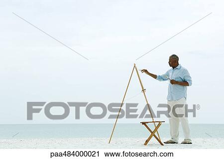 Stock Image - Senior man painting canvas at the beach, side view. Fotosearch