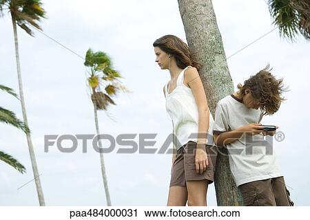 Stock Image - Siblings leaning against tree trunk, teen girl listening to earphones, boy playing handheld video game. Fotosearch