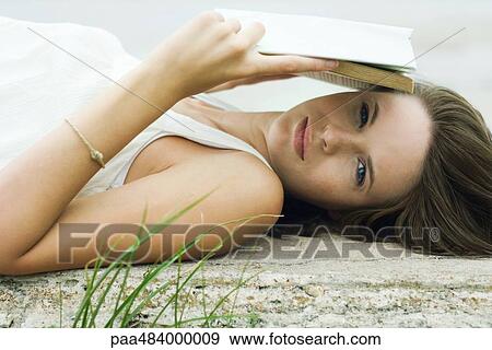Stock Photo - Teenage girl lying on the ground, holding book over face, looking at camera. Fotosearch