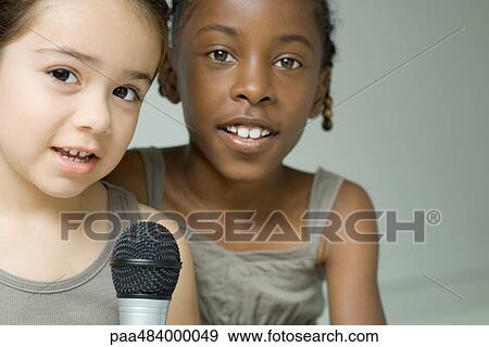 Two young girls singing into microphone together, both looking at camera View Large Photo Image Stock Photo - Two young girls singing into microphone together, both looking at camera. Fotosearch