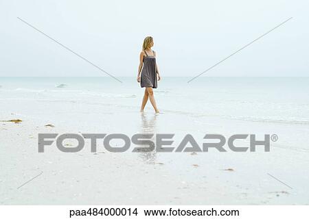 Picture - Woman in sundress walking along beach, looking at view. Fotosearch