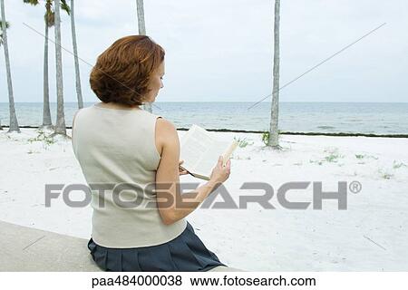 Stock Photo - Woman sitting at the beach reading book, rear view. Fotosearch