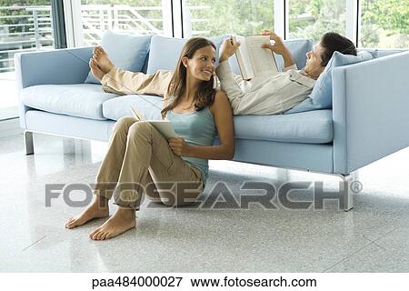 Young man lying on couch reading book, girlfriend sitting on the ground next to him View Large Photo Image Stock Photo - Young man lying on couch reading book, girlfriend sitting on the ground next to him. Fotosearch