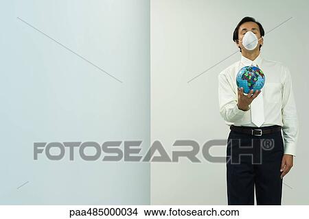 Picture - Businessman wearing pollution mask, holding out globe, eyes closed, front view. Fotosearch