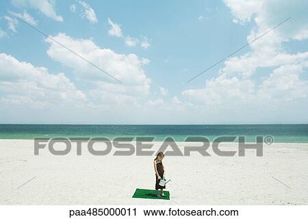 Stock Image - Girl standing on square of artificial turf on beach, holding watering can, high angle view. Fotosearch
