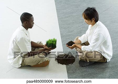 Stock Photo - Two males sitting face to face, one holding out grass, the other holding out soil, side view. Fotosearch