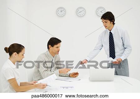 Stock Photo - Businessman discussing document with colleagues, clocks on wall in background. Fotosearch