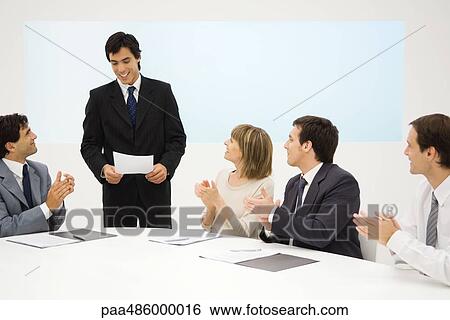 Businessman standing, holding document, seated colleagues clapping, smiling View Large Photo Image Stock Photograph - Businessman standing, holding document, seated colleagues clapping, smiling. Fotosearch