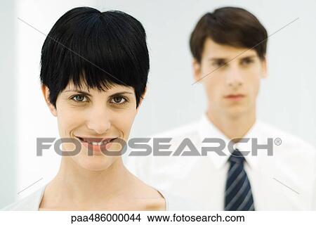 Businesswoman smiling at camera, close-up, male colleague in background View Large Photo Image Picture - Businesswoman smiling at camera, close-up, male colleague in background. Fotosearch