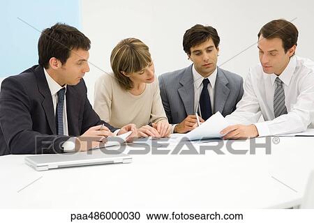 Four business partners sitting together at table, looking at document View Large Photo Image Stock Image - Four business partners sitting together at table, looking at document. Fotosearch