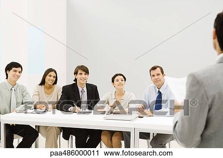 Stock Image - Group of professionals sitting at table, smiling, looking at man standing in foreground. Fotosearch