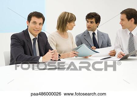 Stock Photograph - Team of business associates sitting at table, discussing document, one smiling at camera. Fotosearch