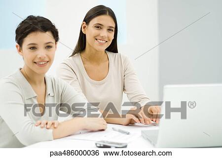 Two businesswomen sitting at desk together next to laptop computer, one smiling at camera View Large Photo Image Stock Photograph - Two businesswomen sitting at desk together next to laptop computer, one smiling at camera. Fotosearch