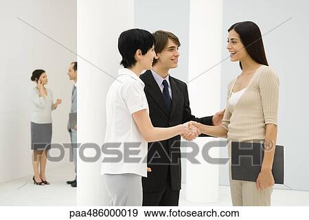 Two businesswomen standing beside male colleague, shaking hands, smiling at each other View Large Photo Image Stock Photo - Two businesswomen standing beside male colleague, shaking hands, smiling at each other. Fotosearch