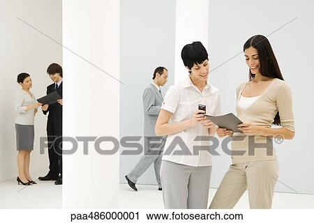 Two businesswomen standing together, looking down at folder, one holding cell phone, coworkers in background View Large Photo Image Stock Image - Two businesswomen standing together, looking down at folder, one holding cell phone, coworkers in background. Fotosearch