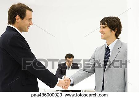Stock Photo - Two male professionals shaking hands, smiling at each other, colleague seated in background. Fotosearch
