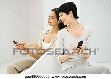 Two women sitting together, both holding cell phones, one looking over shoulder, the other pointing View Large Photo Image Stock Photograph - Two women sitting together, both holding cell phones, one looking over shoulder, the other pointing. Fotosearch