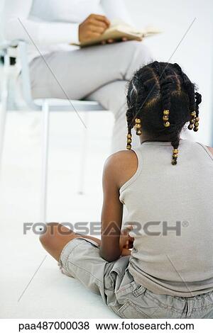 Stock Photo - Girl sitting on floor, listening to mother reading book, rear view, cropped. Fotosearch