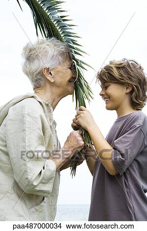 Grandmother and grandson face to face, holding palm leaf together, side view View Large Photo Image Picture - Grandmother and grandson face to face, holding palm leaf together, side view. Fotosearch