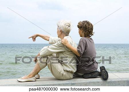 Grandmother and grandson sitting together by the sea, woman pointing, rear view View Large Photo Image Stock Image - Grandmother and grandson sitting together by the sea, woman pointing, rear view. Fotosearch