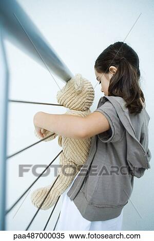 Stock Photography - Little girl holding teddy bear, leaning against railing, side view. Fotosearch