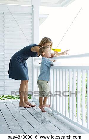 Stock Photograph - Mother and son standing on porch together, woman holding binoculars and pointing, both looking away. Fotosearch