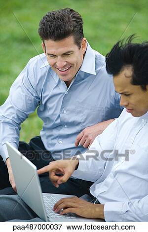 Stock Photo - Two businessmen sitting side by side outdoors, both looking down at laptop computer. Fotosearch