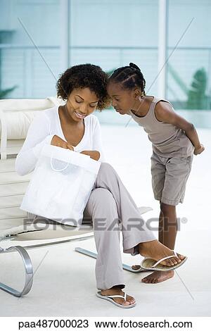 Woman sitting in chair, holding gift bag, daughter standing beside her, both looking down View Large Photo Image Stock Image - Woman sitting in chair, holding gift bag, daughter standing beside her, both looking down. Fotosearch