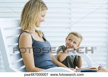 Woman sitting in chair next to son, holding book, both smiling at each other View Large Photo Image Stock Photo - Woman sitting in chair next to son, holding book, both smiling at each other. Fotosearch