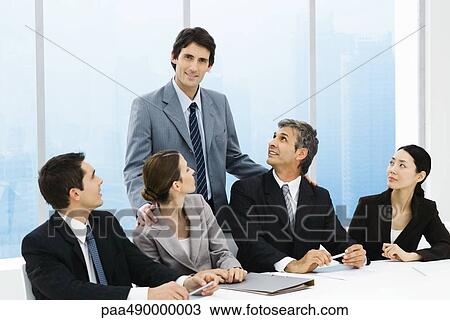 Stock Image - Business leader standing behind group of colleagues at meeting table, smiling at camera. Fotosearch
