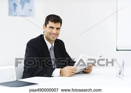 Businessman sitting at desk in office, reading newspaper, smiling at camera View Large Photo Image Stock Photo - Businessman sitting at desk in office, reading newspaper, smiling at camera. Fotosearch
