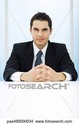 Businessman sitting at table with hands clasped, looking at camera View Large Photo Image Picture - Businessman sitting at table with hands clasped, looking at camera. Fotosearch
