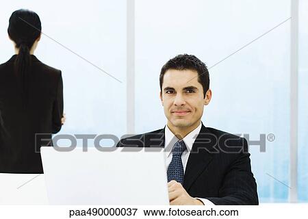 Stock Photo - Businessman sitting behind computer screen, smiling at camera. Fotosearch
