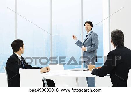 Stock Photo - Businessmen in meeting, one standing, pointing out window. Fotosearch