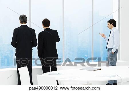 Businessmen standing by window in conference room, one checking cell phone View Large Photo Image Stock Image - Businessmen standing by window in conference room, one checking cell phone. Fotosearch