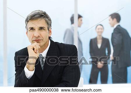 Graying businessman with hand under chin, looking away, colleagues chatting in background View Large Photo Image Stock Photo - Graying businessman with hand under chin, looking away, colleagues chatting in background. Fotosearch