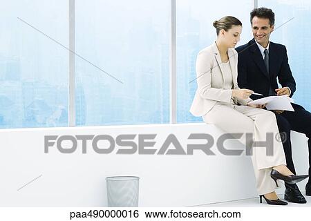 Male and female executives sitting on ledge by window, discussing document View Large Photo Image Stock Photograph - Male and female executives sitting on ledge by window, discussing document. Fotosearch