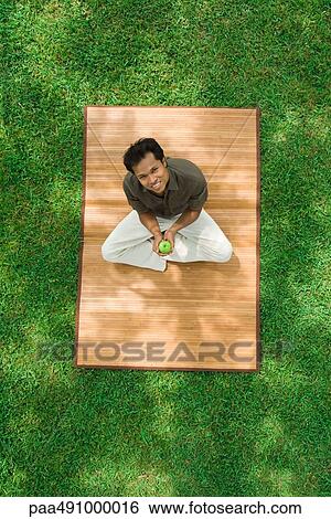 Man sitting on the ground outdoors, holding apple, smiling at camera, high angle view View Large Photo Image Stock Photograph - Man sitting on the ground outdoors, holding apple, smiling at camera, high angle view. Fotosearch