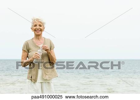 Stock Photography - Senior woman standing at the beach, holding bottle of water, smiling at camera. Fotosearch